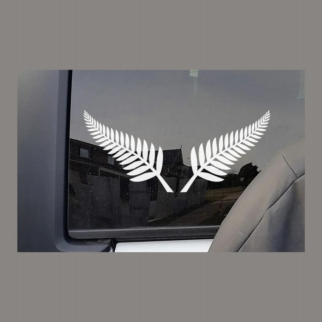 Silver fern decal pair – two white fern fronds on a car side window with houses reflected in the glass
